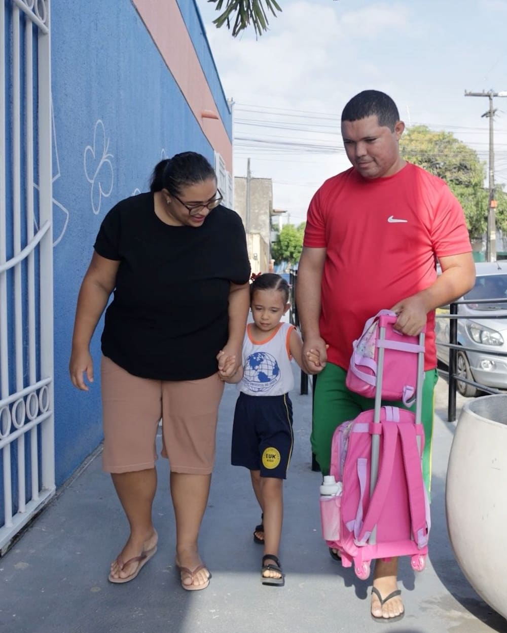Mãe e pai chegando ao colégio de mãos dadas com o filho, que leva a mochila
